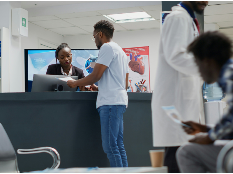 A imagem mostra o interior de uma clínica médica ou hospital. No centro da imagem, um homem está em pé diante de uma recepcionista, aparentemente fazendo um cadastro ou entrega de documentos. A recepcionista está atrás de um balcão, olhando atenta para os papéis que ele entrega.