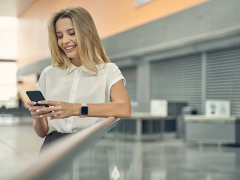 A imagem mostra uma mulher sorridente em um ambiente moderno e corporativo, usando um smartphone. Ela parece estar interagindo com um aplicativo, o que transmite praticidade, conectividade e satisfação com a experiência digital. Ela veste roupas sociais e usa um smartwatch, reforçando o perfil de alguém que valoriza eficiência e tecnologia no dia a dia.