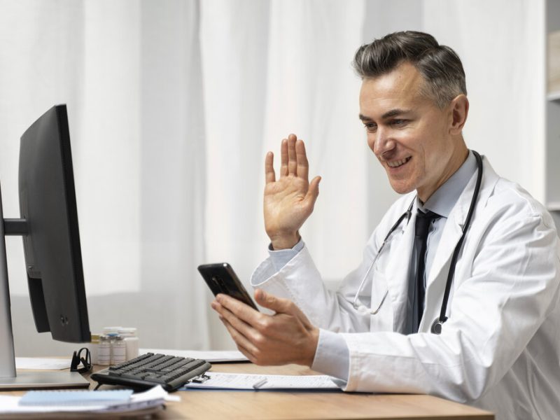Médico de jaleco branco, com estetoscópio ao redor do pescoço, realizando um teleatendimento com um paciente.