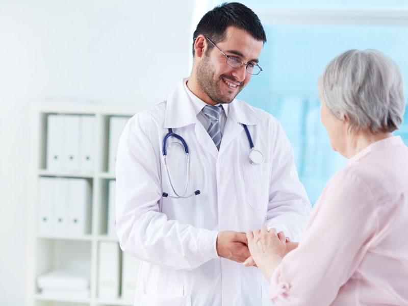 RQE médico: médico sorrindo e segurando a mão de uma idosa em um consultório.
