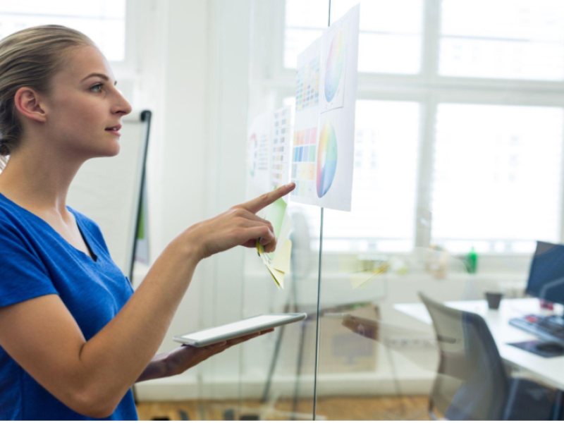 A imagem mostra uma mulher em um ambiente corporativo ou de escritório moderno, de perfil, interagindo com um painel de vidro transparente onde estão fixados gráficos e diagramas coloridos, como gráficos de pizza e tabelas.