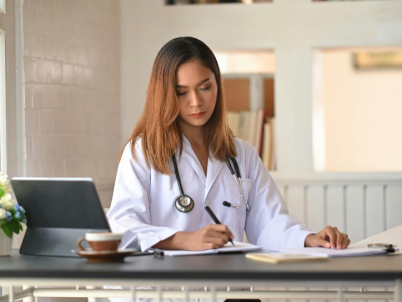 A imagem mostra uma profissional da área da saúde, possivelmente uma médica, sentada em uma mesa enquanto escreve em um caderno ou ficha.