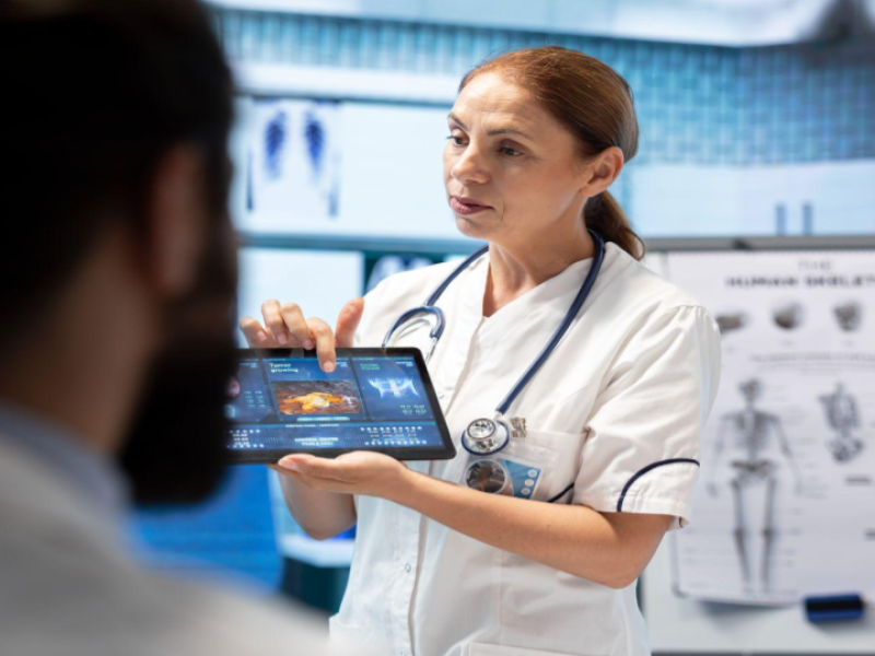 A imagem mostra uma profissional de saúde, provavelmente uma médica ou enfermeira, usando uniforme branco e estetoscópio no pescoço, explicando informações a um paciente em um ambiente clínico.
