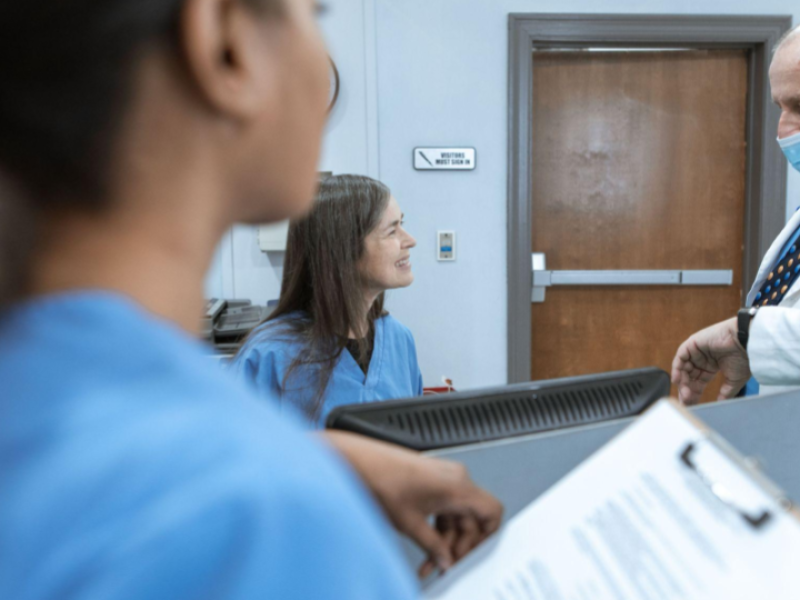A imagem mostra um ambiente médico ou hospitalar, focado na interação entre profissionais de saúde em um balcão ou recepção.