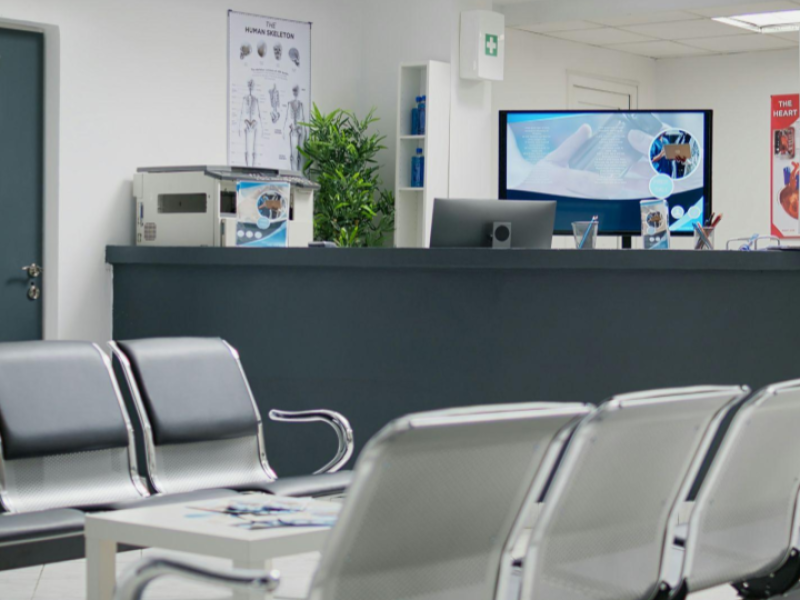 Esta imagem mostra a recepção e sala de espera de um centro médico ou clínica de saúde, moderna e organizada, no momento sem pacientes ou atendentes.
