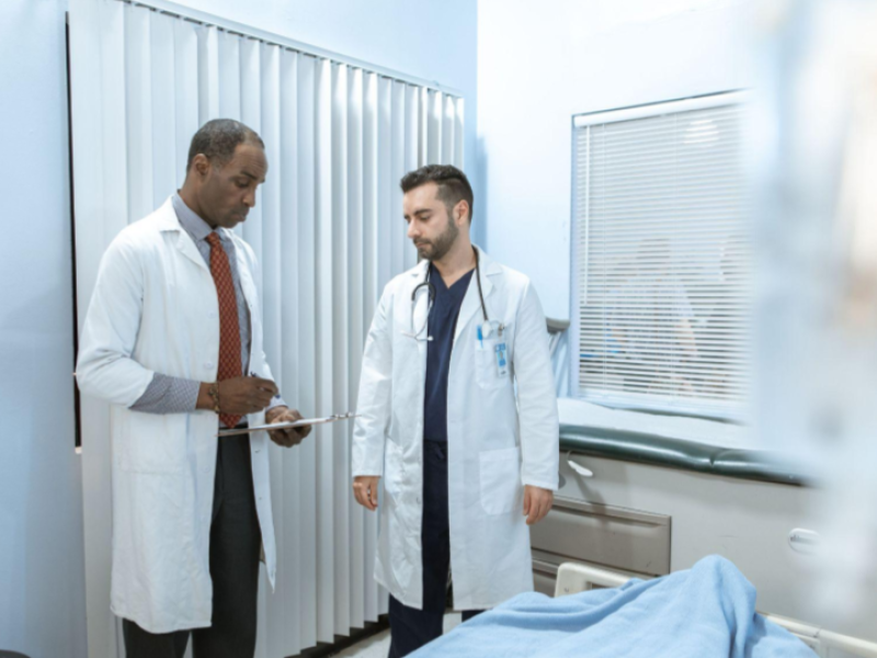 A imagem retrata dois médicos conversando no que parece ser um quarto de hospital ou uma sala de exames clínicos. O ambiente transmite uma atmosfera profissional, colaborativa e de cuidado médico.