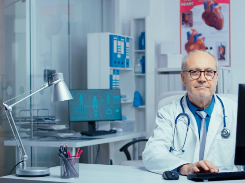 A imagem mostra um médico sentado em seu consultório, trabalhando em frente a um computador. Ele usa jaleco branco, estetoscópio no pescoço e óculos, e olha para a câmera com uma expressão calma e profissional, transmitindo confiança e experiência.