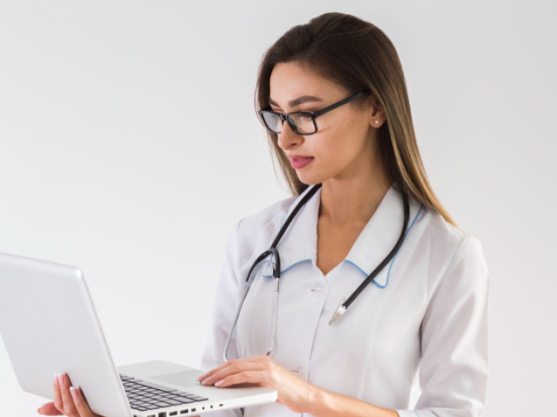 A imagem mostra uma profissional da área da saúde, vestida com jaleco branco e estetoscópio no pescoço, utilizando um notebook.