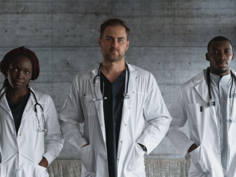 A imagem mostra um retrato de um grupo diversificado composto por três profissionais da saúde (médicos ou enfermeiros), posando de forma confiante e séria.