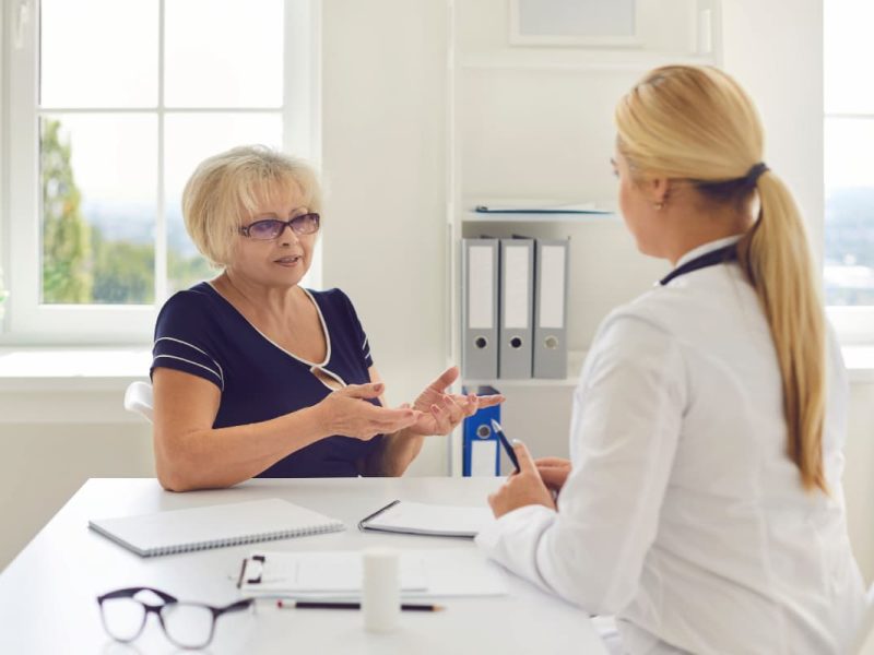 Paciente idosa em consulta de acompanhamento médico, conversando com uma profissional de saúde em um consultório iluminado.