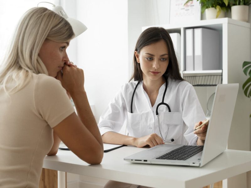 médica de jaleco branco faz uma anamnese completa junto com uma paciente, utilizando um notebook