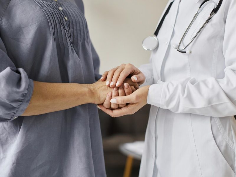 Médica, de jaleco branco, segurando as duas mãos do paciente de roupa cinza.