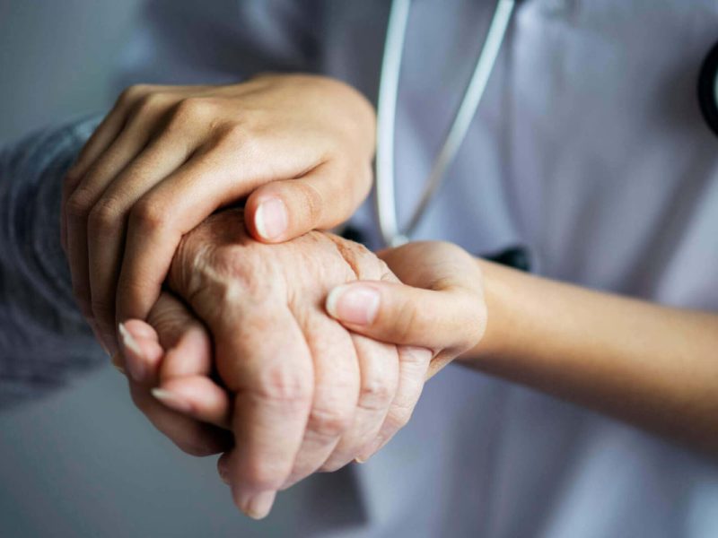 Mãos de profissional de saúde segurando a mão de um paciente idoso, simbolizando o atendimento preferencial na telemedicina.