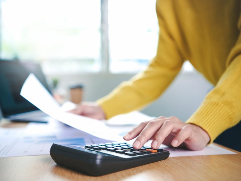Pessoa utilizando calculadora para gerenciar cobrança recorrente, com papéis e laptop ao fundo.