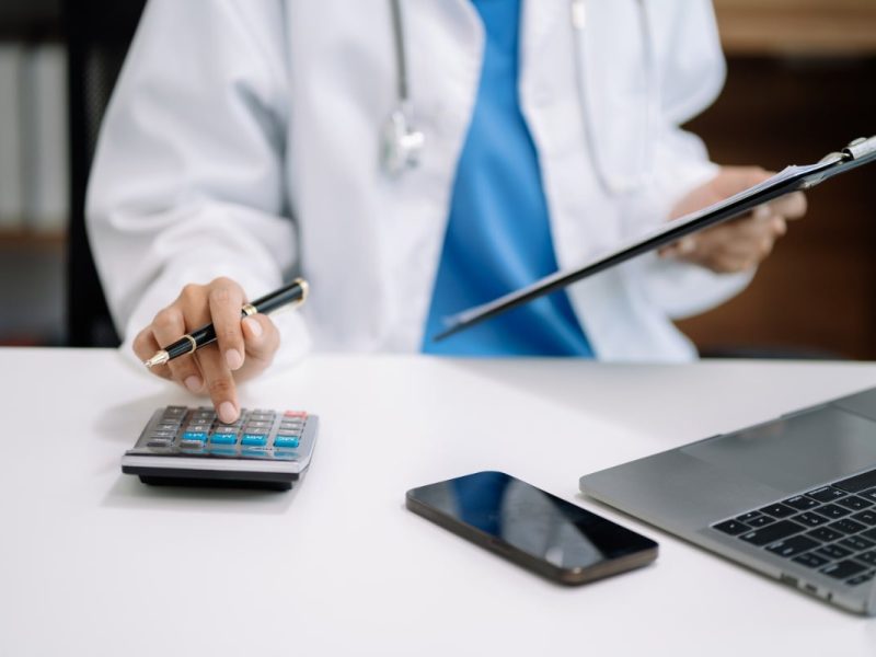 Médico no seu consultório com uma calculadora, notebook e celular na mesa, aprendendo a como calcular o pró-labore médico.