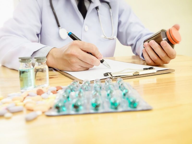 Um médico de jaleco branco usa uma caneta e sabe como preencher receita de rifocina de forma correta, com medicamos ao lado na mesa