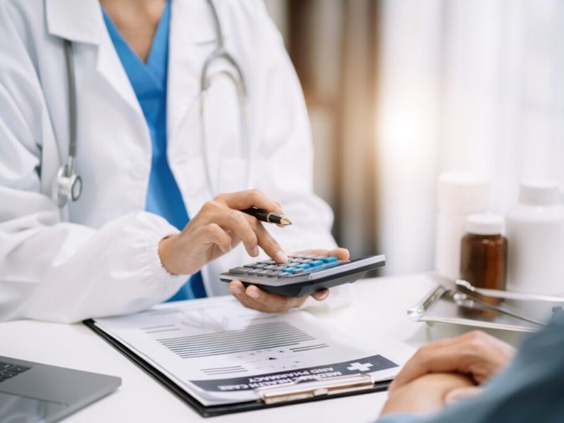 Uma médica com jaleco branco e estetoscópio utiliza uma calculadora para fazer a contabilidade da clínica