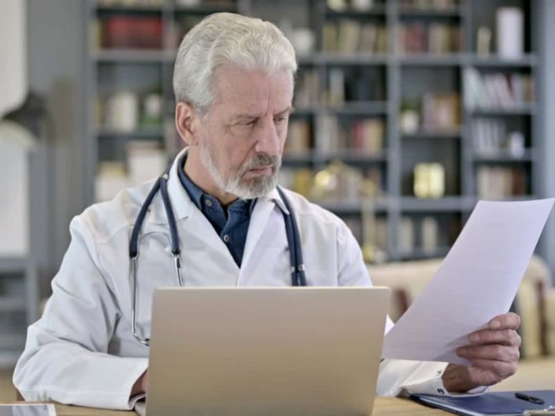 Médico olhando prontuário eletrônico do paciente (PEP) em papel com um computador na frente.