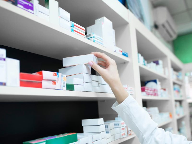 Profissional de saúde realizando a dispensação de medicamentos em uma farmácia, organizando caixas de remédios na prateleira.