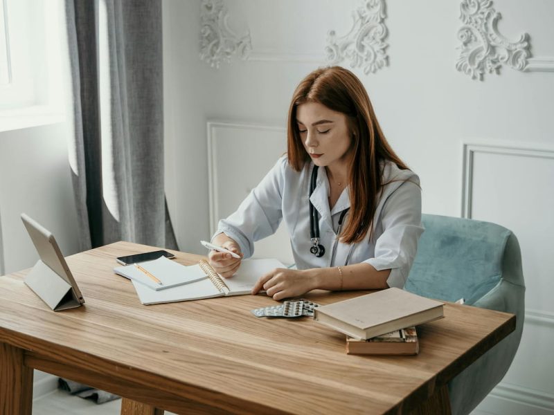 Médica preenchendo a DMED em um consultório, com laptop, papéis e estetoscópio sobre a mesa.