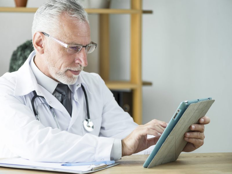 Esta imagem apresenta um homem, um médico sênior, com cabelo e barba grisalhos, com cerca de 60 anos. Ele está sentado em uma mesa de madeira, concentrado em seu trabalho.