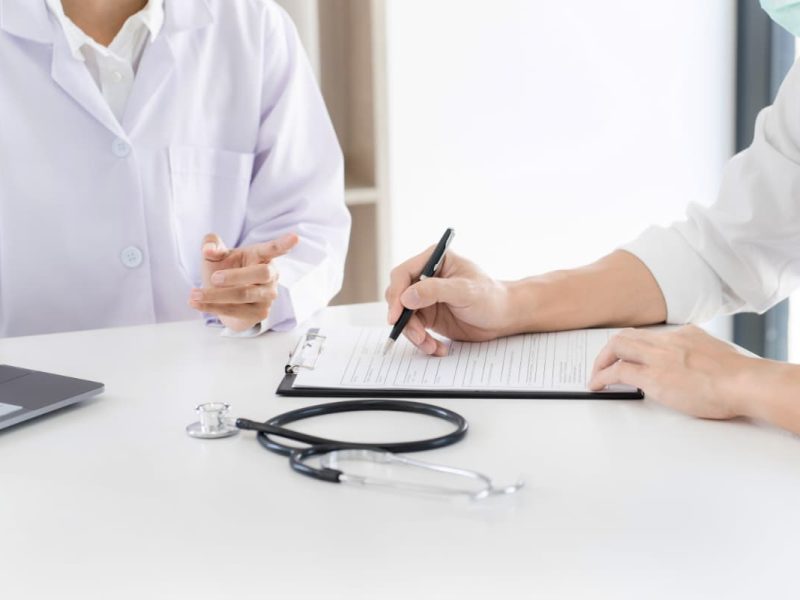 Um profissional de saúde com jaleco branco está preenchendo um documento de encaminhamento médico ao lado de um paciente.