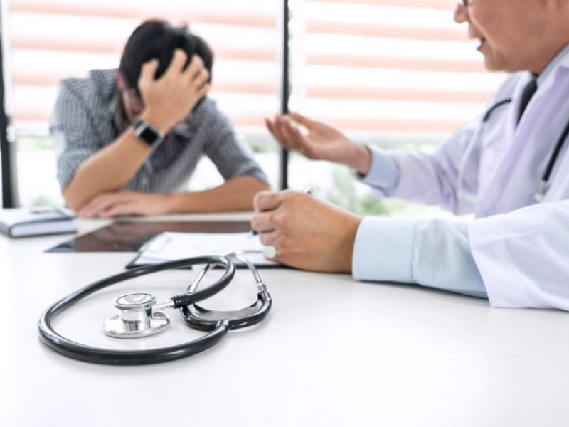 Paciente angustiado com médico, estetoscópio na mesa, sugere erro médico.