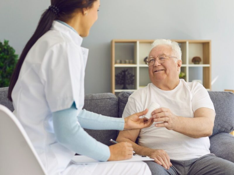 Um idoso sorridente recebe seus medicamentos de uma profissional de saúde em casa, representando o conceito de home care