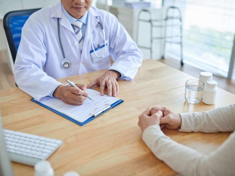 Médico discutindo inovações em saúde com paciente em clínica, com equipamentos médicos e documentos na mesa.