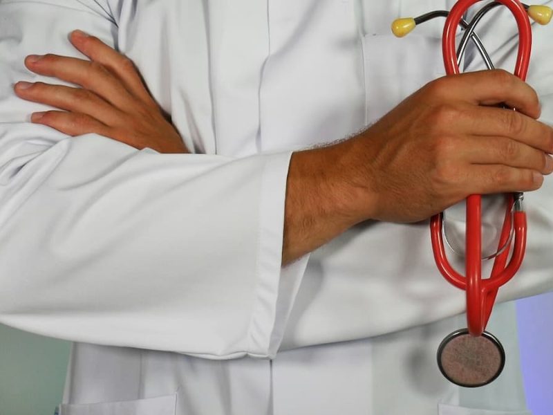 Fotografia de um homem, vestindo uma bata de médico, de braços cruzados.