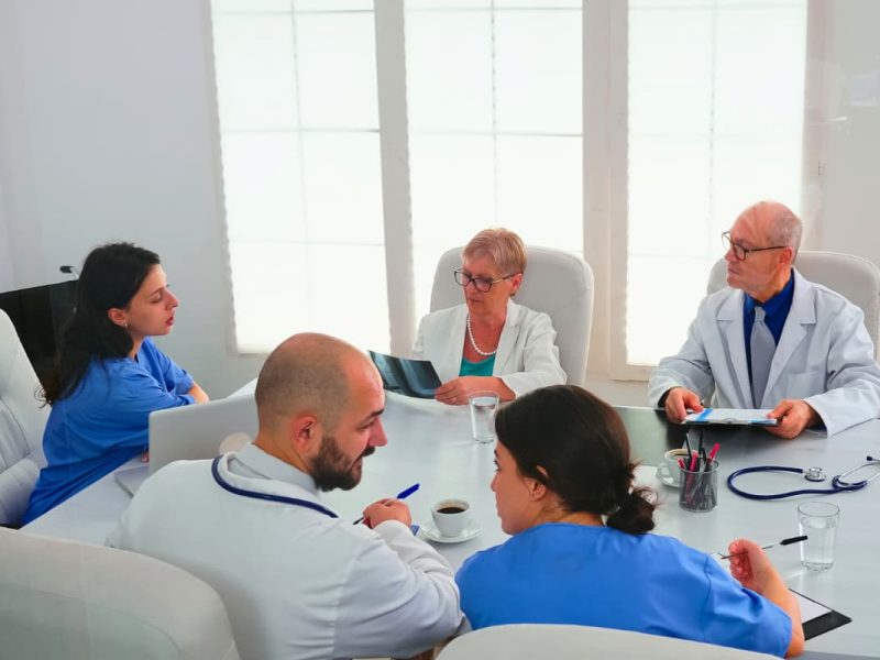 Reunião de junta médica com profissionais analisando exames e discutindo casos clínicos em uma sala de conferência.