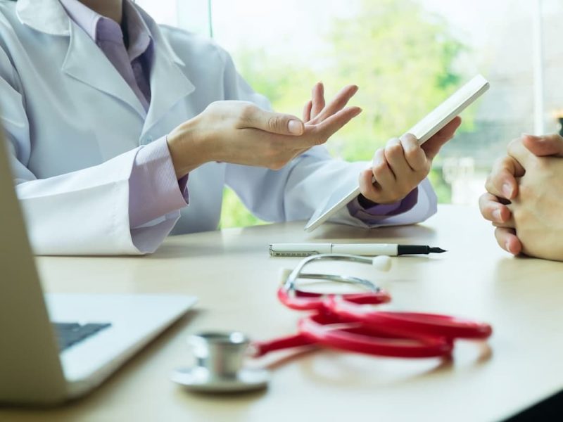 Médico explicando pagamento online para paciente via tablet, com estetoscópio e laptop ao lado.