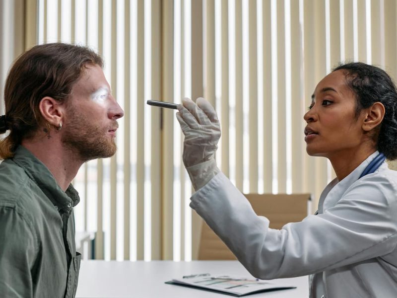 Em plantão médico, um profissional de saúde com jaleco branco examina os olhos de um paciente com uma lanterna clínica. Seguindo procedimentos do triple aim