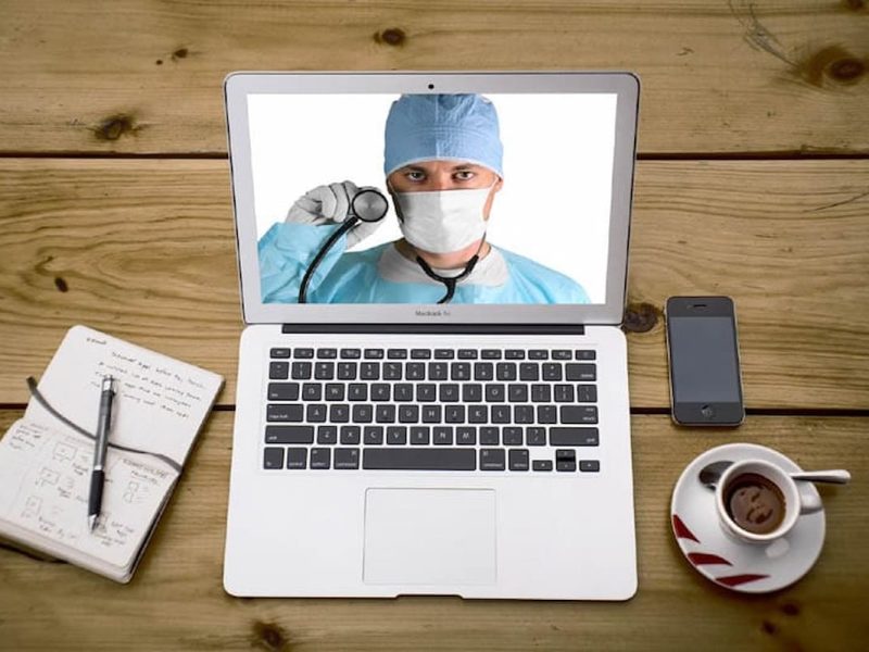 Médico em tela de laptop, simbolizando plataforma de telemedicina, com estetoscópio, caderno e celular ao lado.