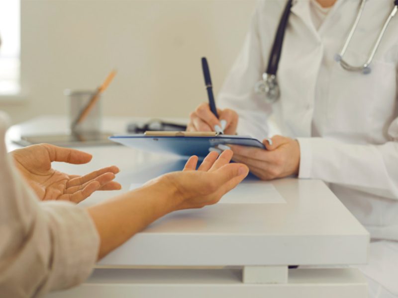 Uma médica de jaleco branco e estetoscópio preenche uma receita amarela para uma paciente, que está sentada na sua frente
