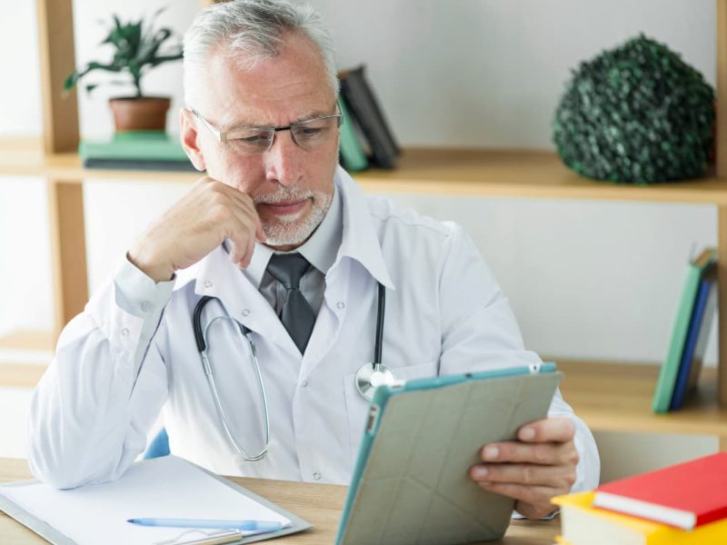 Homem de jaleco branco sentado à mesa, utilizando um tablet, representando saúde financeira para clínicas médicas.