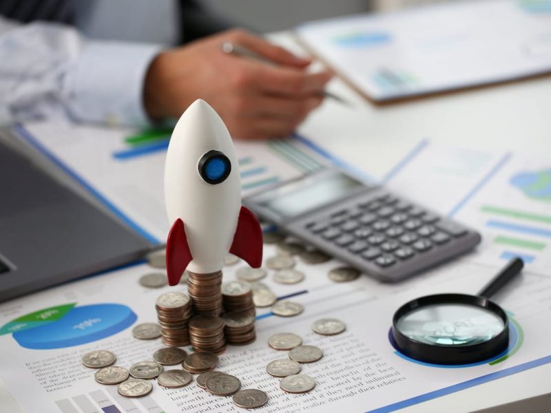 Modelo de foguete sobre pilhas de moedas, gráficos financeiros ao fundo, simbolizando sistema de controle financeiro.