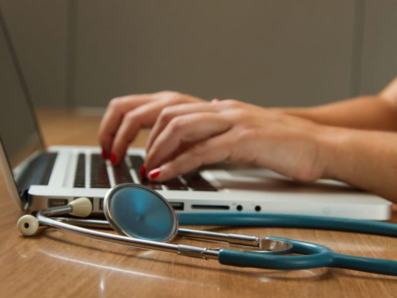Mãos digitando em laptop com estetoscópio ao lado, representando sistema para laudos médicos online.