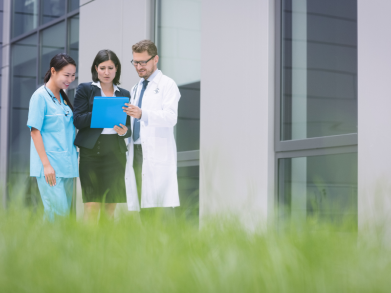 A imagem mostra três profissionais conversando em um ambiente externo de um prédio corporativo ou hospitalar, transmitindo colaboração e alinhamento entre equipes da área da saúde.