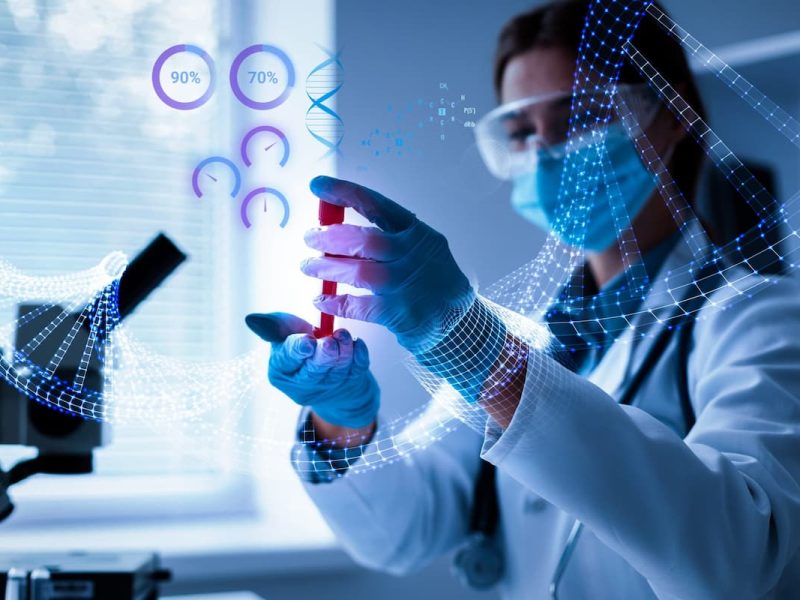 Biomédica com jaleco branco, máscara e óculos de proteção. Na frente dela, existe um microscópio com a tecnologia na saúde.