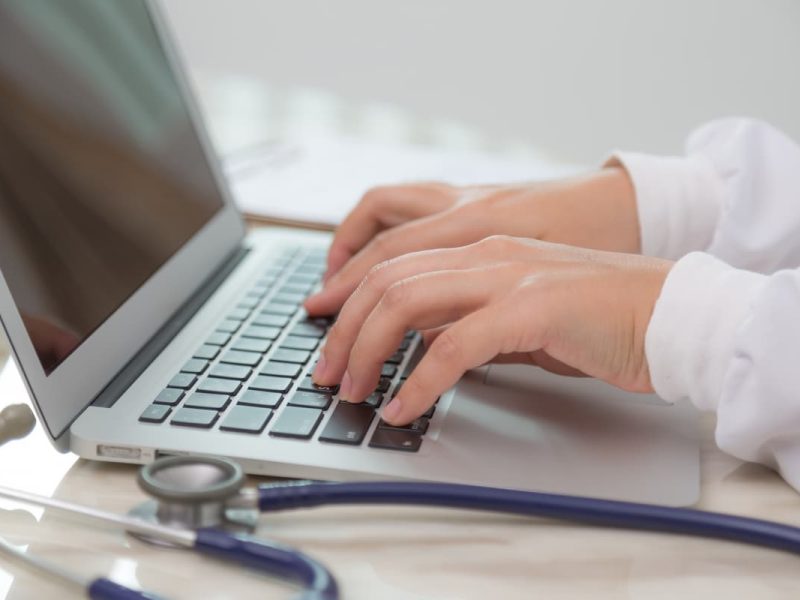 Médico digitando em laptop com estetoscópio ao lado, representando diferentes tipos de prontuário eletrônico na saúde.
